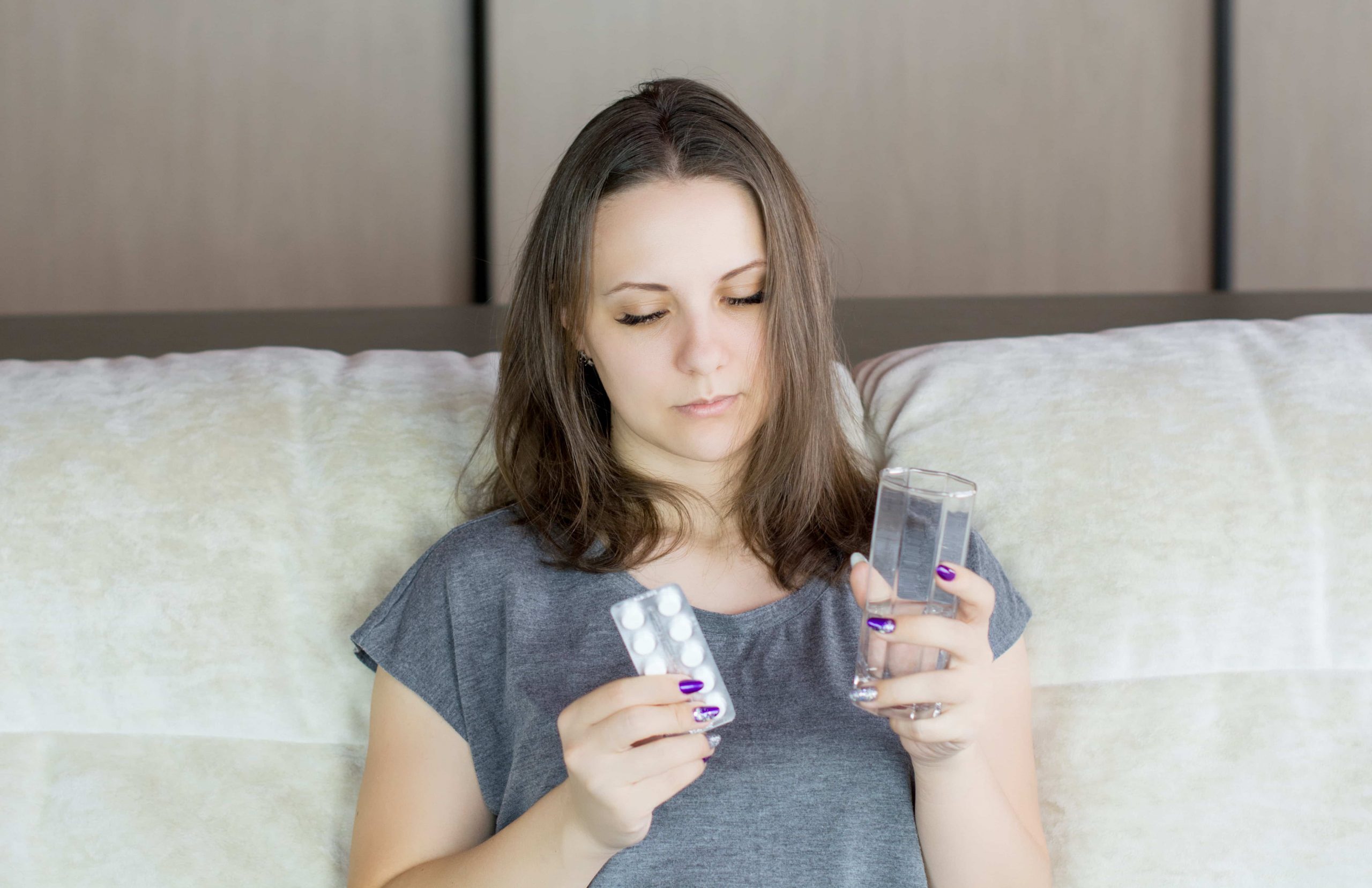 إمرأة شابة تجلس على السرير، تحمل كوب ماء ممتليء للنصف وشريط دواء وتنظر إليهما